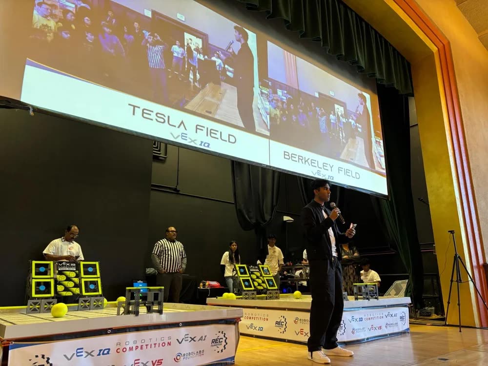 Arya Sasikumar on stage with a microphone at the NorCal state championship, the Tesla Field and Berkeley Field scoreboard behind him
