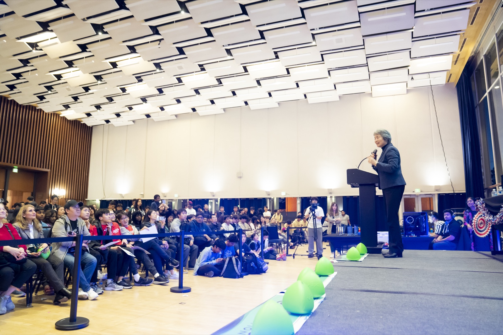 Wide view of the packed Pauley Ballroom with a speaker at the podium and game elements on the floor