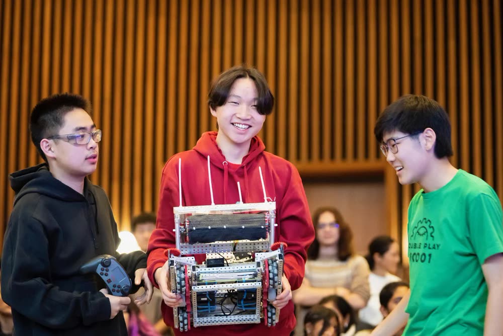 Three students laughing, one carrying their robot off the field